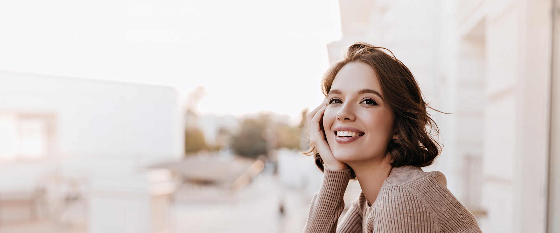 The image features a woman with her head tilted slightly to the side, smiling softly, sitting outdoors, wearing a light-colored top, with a blurred background that suggests an urban setting.