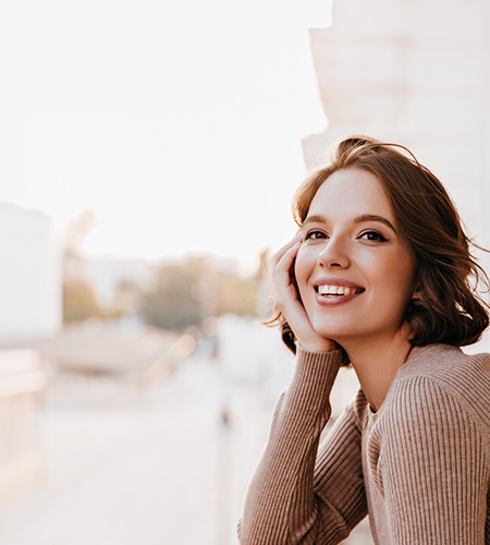 A woman with short hair smiling at the camera while leaning on her hand, set against a blurred cityscape background during sunset.