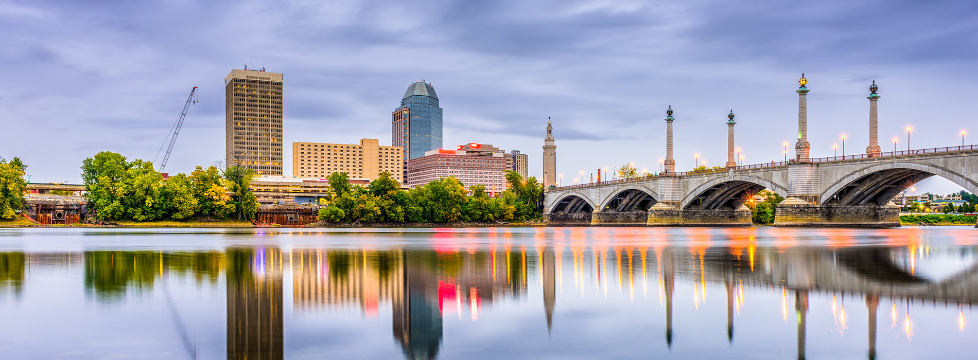 The image shows a city skyline at night with a bridge over a river reflecting lights on its surface, and a reflection of the bridge and cityscape on the water s surface.