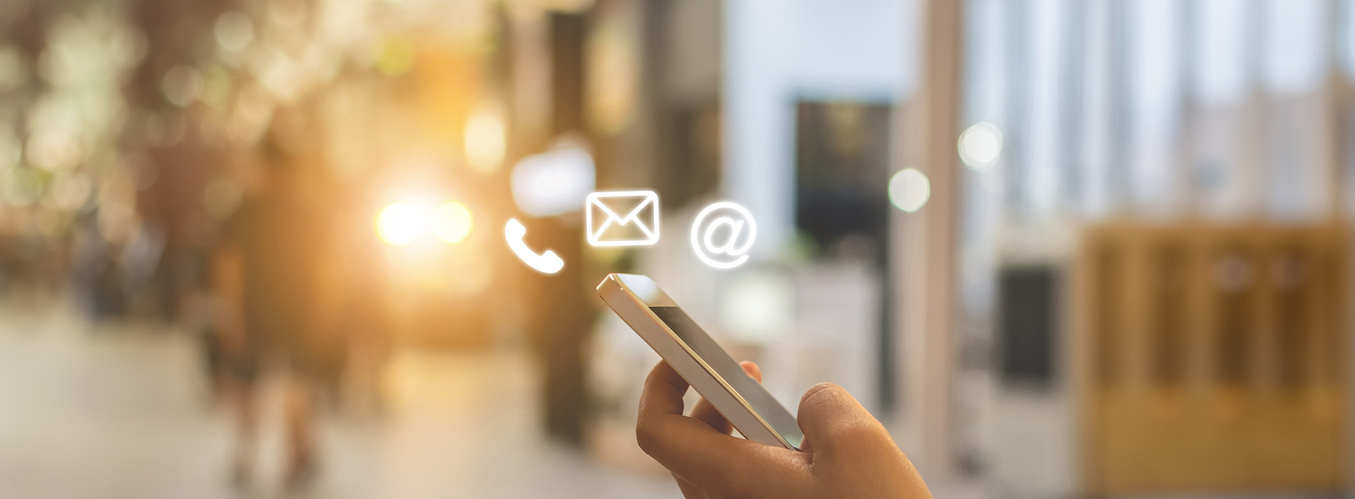 The image shows a smartphone with an email icon on its screen being held by a person s hand, which is visible at the bottom of the frame. The background appears to be an indoor setting with blurred lights and a dimly lit environment that suggests a public space, possibly a shopping center or airport lounge.