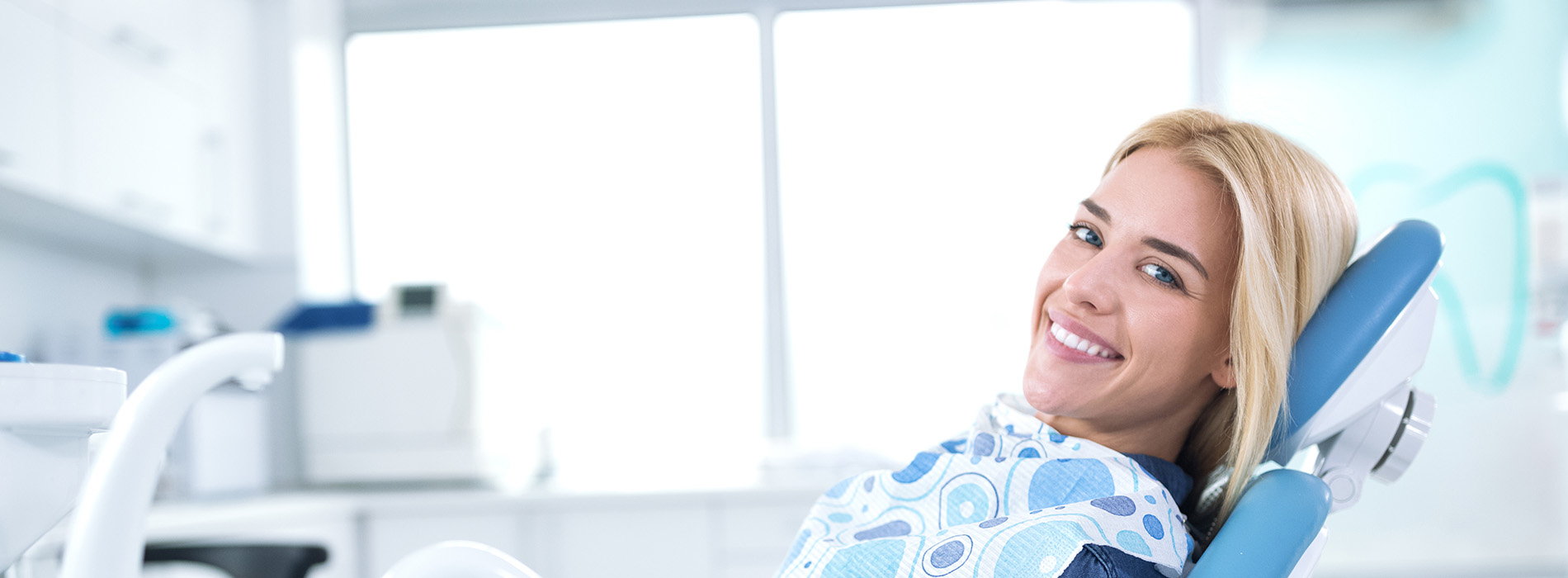 A smiling woman sitting in an orthodontist s chair with dental equipment in the background.