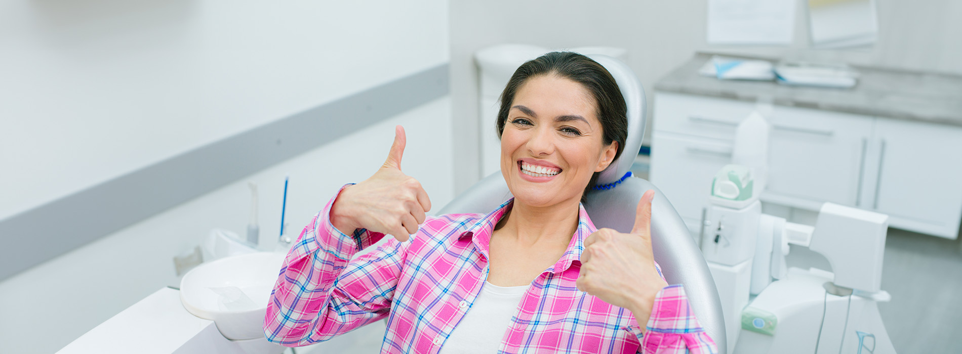 A woman sitting in a dental chair giving a thumbs up gesture with a big smile, wearing a blue plaid shirt.
