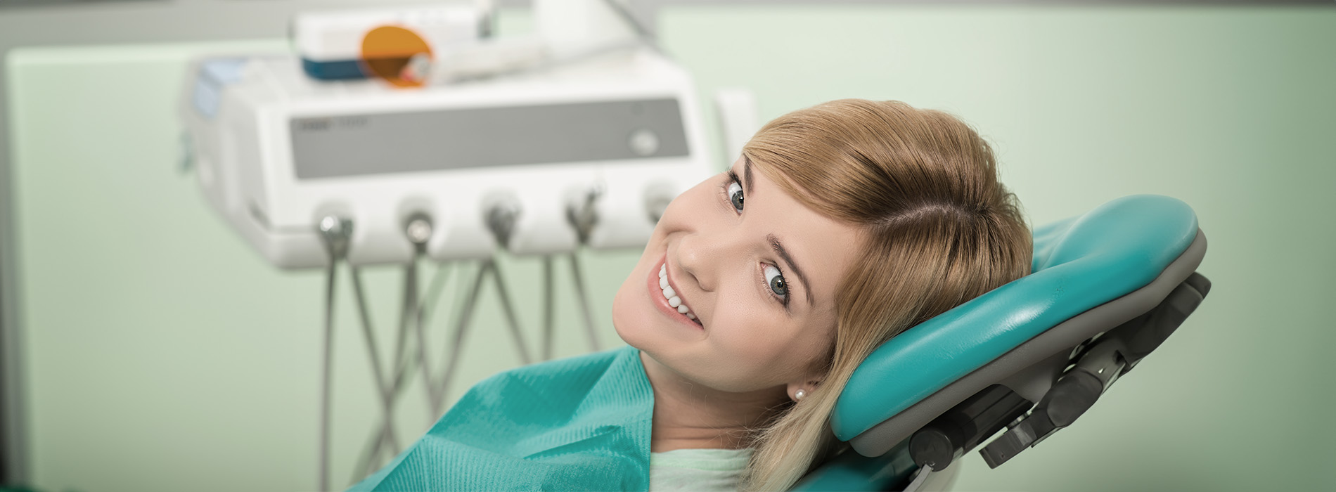 The image features a woman sitting in a dental chair, smiling at the camera with her eyes closed, wearing a blue shirt, with dental equipment visible behind her.