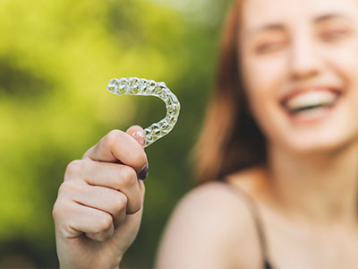 The image features a person holding an oversized toothbrush with a smile on their face, set against a blurred background.
