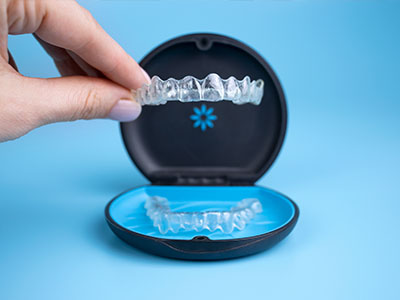 The image shows a hand holding an open box containing a set of clear braces with transparent wire elements, placed on a blue surface against a blurred background.
