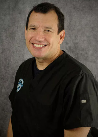 The image features a man wearing a white lab coat with a logo on the left chest area, standing against a grey background with a black and white pattern, smiling at the camera.
