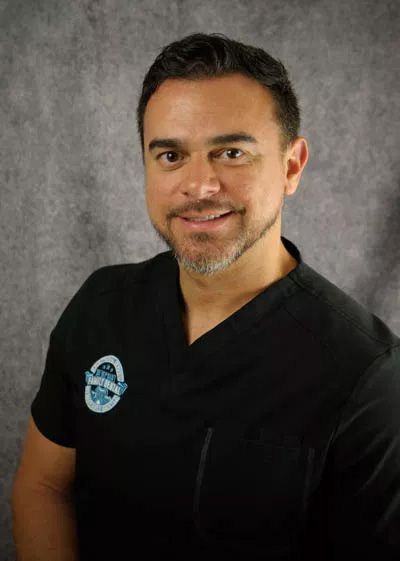 A man wearing a black shirt with a stethoscope around his neck, smiling at the camera, against a backdrop with a gray wall.
