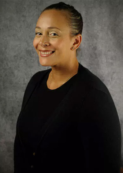 The image features a smiling woman with short hair, wearing a black top, posing for a portrait against a dark background.