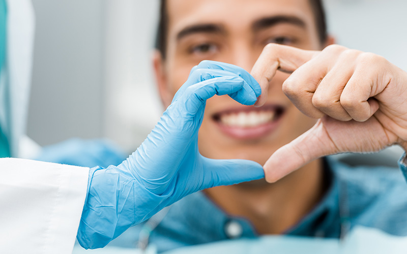 A person wearing blue gloves holds up a heart shape with both hands while another individual looks on behind a medical mask.