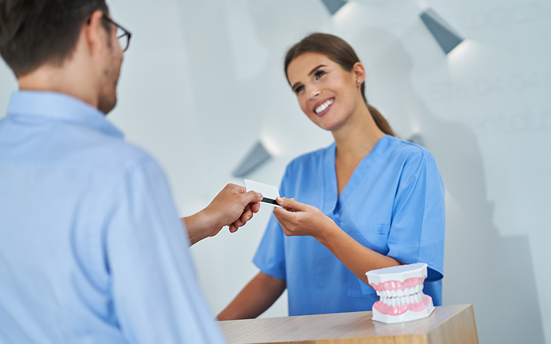 The image depicts two individuals at a dental clinic  one person is handing over a card to another who appears to be a dental professional, both are wearing masks and standing behind a reception desk.