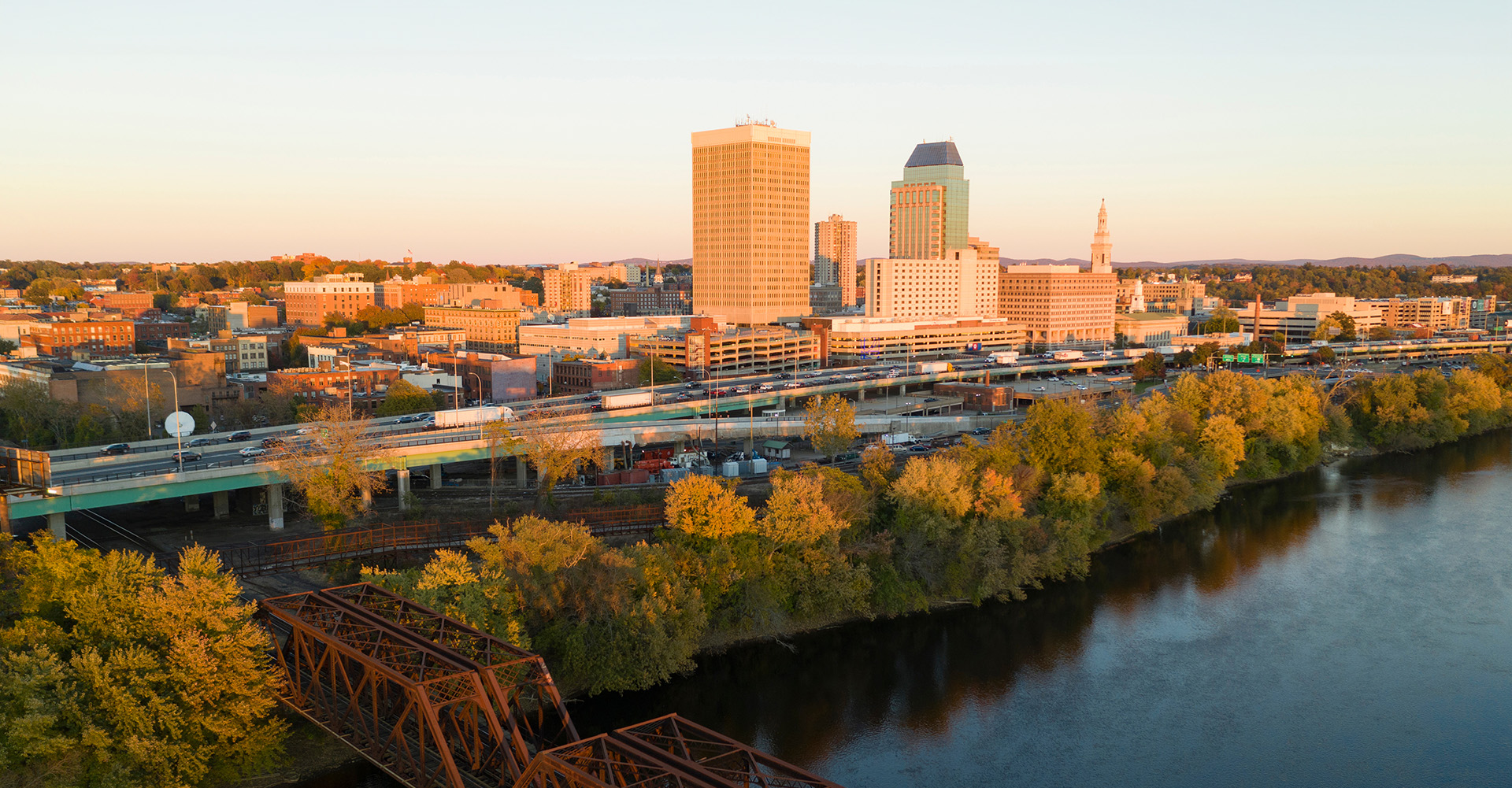 The image depicts a cityscape with a prominent bridge over a river during sunset, featuring a skyline with buildings and a clear view of the horizon.