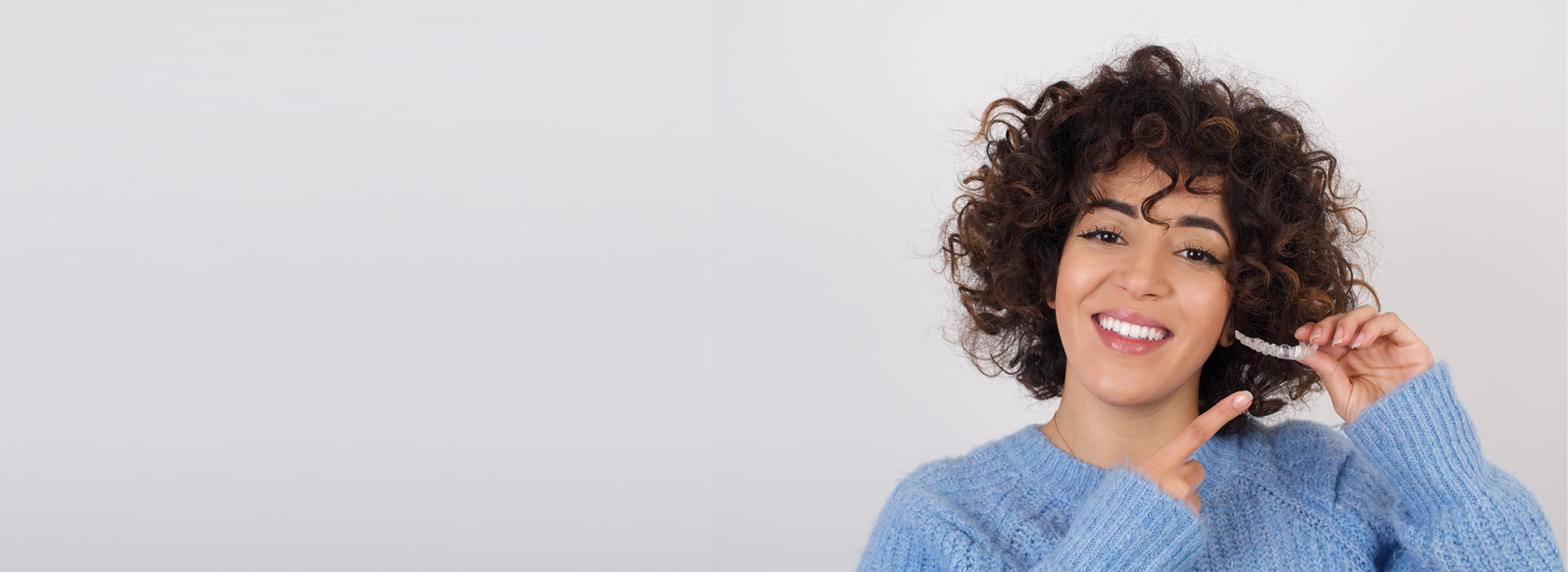 The image features a person with curly hair smiling and holding a toothbrush, set against a plain background with a watermark or logo overlaying the photo.