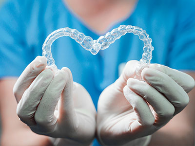 The image depicts a hand holding a clear plastic heart-shaped object with a blue background, which appears to be a transparent representation of a dental impression tray.