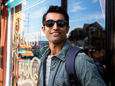 A man wearing sunglasses and a backpack stands outside a storefront with a sign behind him.