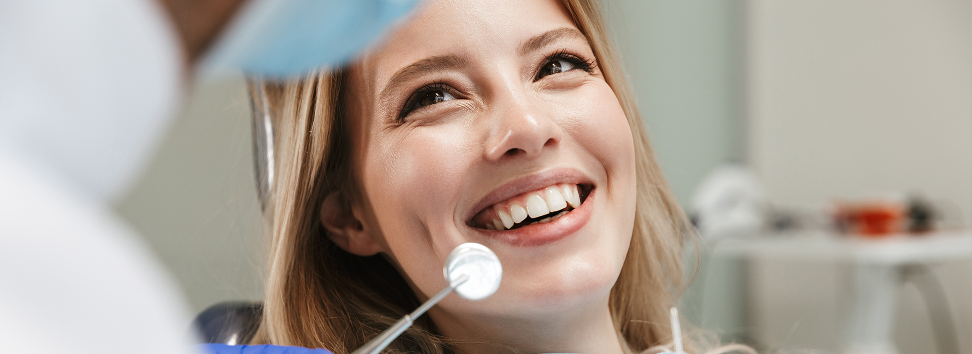 A young woman with a smile is seated at a dental chair, looking towards the camera, while a dentist attends to her, with dental instruments visible in the background.