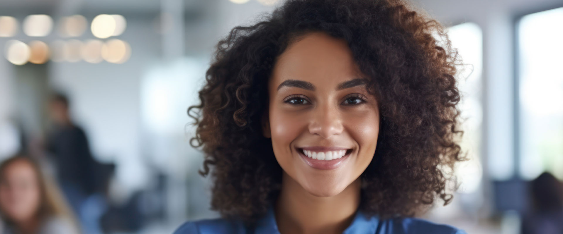 The image shows a smiling woman with curly hair, wearing a blue shirt, standing in an office environment with other people in the background.