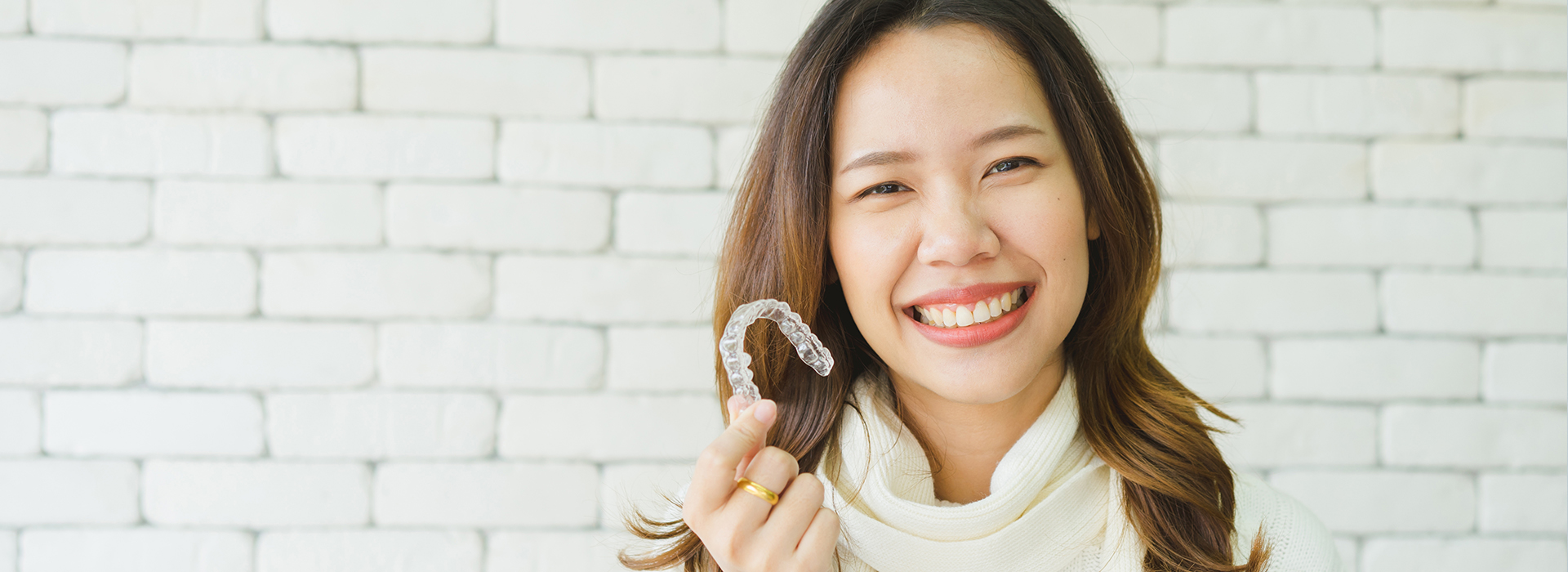 The image shows a person smiling at the camera while holding an object that appears to be a toothbrush, with a blurred background featuring a brick wall.