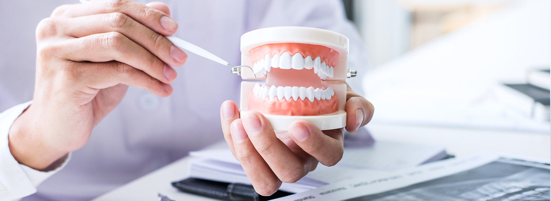 The image shows a person holding a toothbrush with toothpaste and a cup of water near a mouth model, with another person's hand visible above a desk and papers, suggesting an educational or demonstration setting related to dental hygiene.