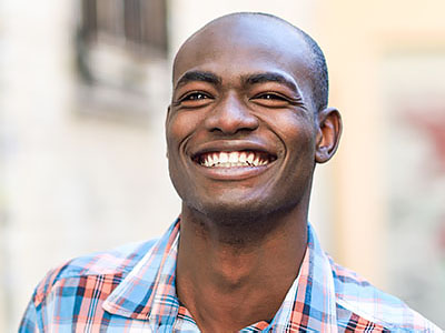 The image shows a smiling man with short hair, wearing a blue plaid shirt and standing against a building background.