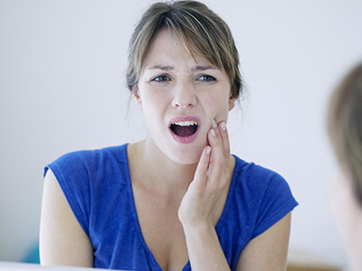 The image shows a woman with her mouth open, displaying teeth, looking at her reflection in a mirror with an expression of concern or surprise.