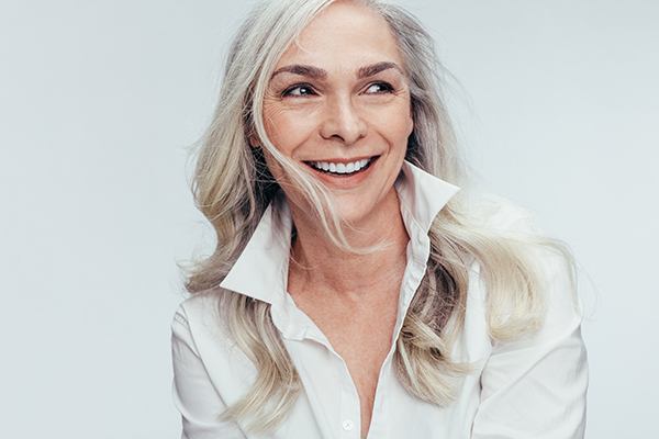 Woman with short hair smiling at camera, wearing a white top and dark pants, set against a plain background.