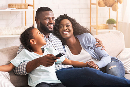 A family of four sitting together on a couch, with a man holding two children while a woman sits behind him. They all appear happy and relaxed, smiling at the camera.