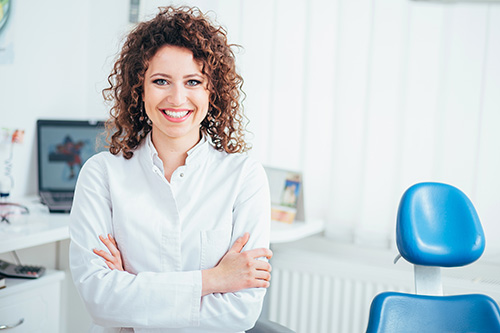 The image shows a woman standing in an office environment with her arms crossed, smiling at the camera. She appears confident and professional, dressed in business attire.