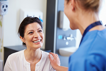 The image shows two people in a medical setting  one woman is seated with a smile, wearing a white shirt, while another person stands behind her, appearing to be engaged in conversation.