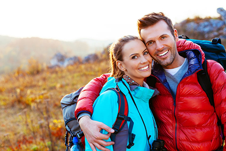 A man and woman posing together outdoors, with the man wearing a backpack and both dressed for cold weather, against a scenic backdrop of autumnal colors.