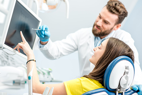 An adult woman with glasses sitting in a dental chair while a dentist in a white coat and blue gloves demonstrates something on a computer screen, both are in a dental office setting.