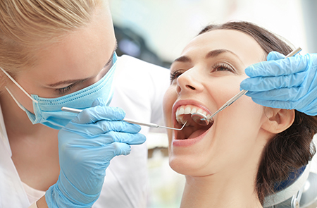 The image features two women in a dental setting  one is seated with her mouth open, receiving dental treatment, while another woman stands by wearing protective gloves and a mask, likely a dental hygienist or dentist, performing the procedure.