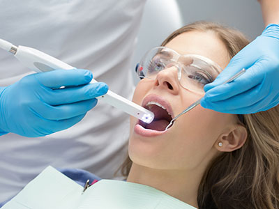 The image shows a woman receiving dental treatment, with a dental professional using a device to examine her mouth while she sits in a dental chair wearing protective blue gloves and a white mask.