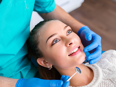 The image shows a smiling woman seated in a dental chair with a dental professional performing a procedure on her mouth.