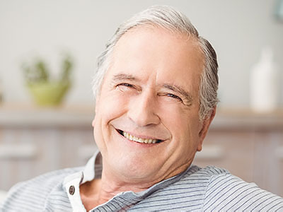 The image shows a smiling older man sitting in a kitchen with his eyes closed and hands resting on his chest.