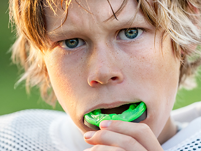The image features a young boy with blonde hair holding a green object in his mouth, possibly a piece of gum or candy, against a blurred background that suggests an outdoor setting, likely during daylight.