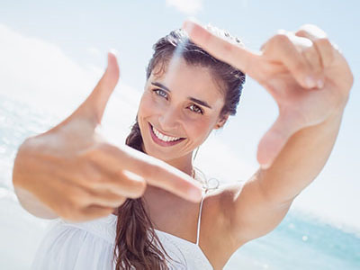 A young woman with long hair smiling at the camera while holding up her hand to frame a shot with her fingers.